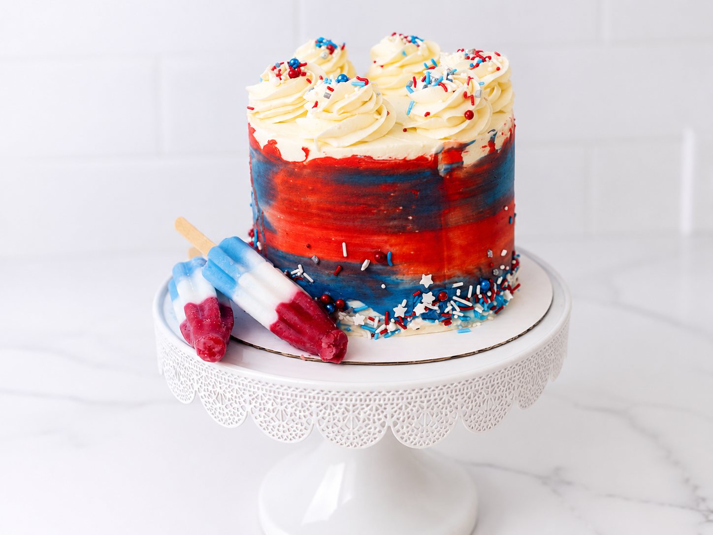 Bomb Pop Cake from Crumb Believable Bakery decorated with red, white, and blue buttercream, patriotic sprinkles, and Bomb Pop popsicles.